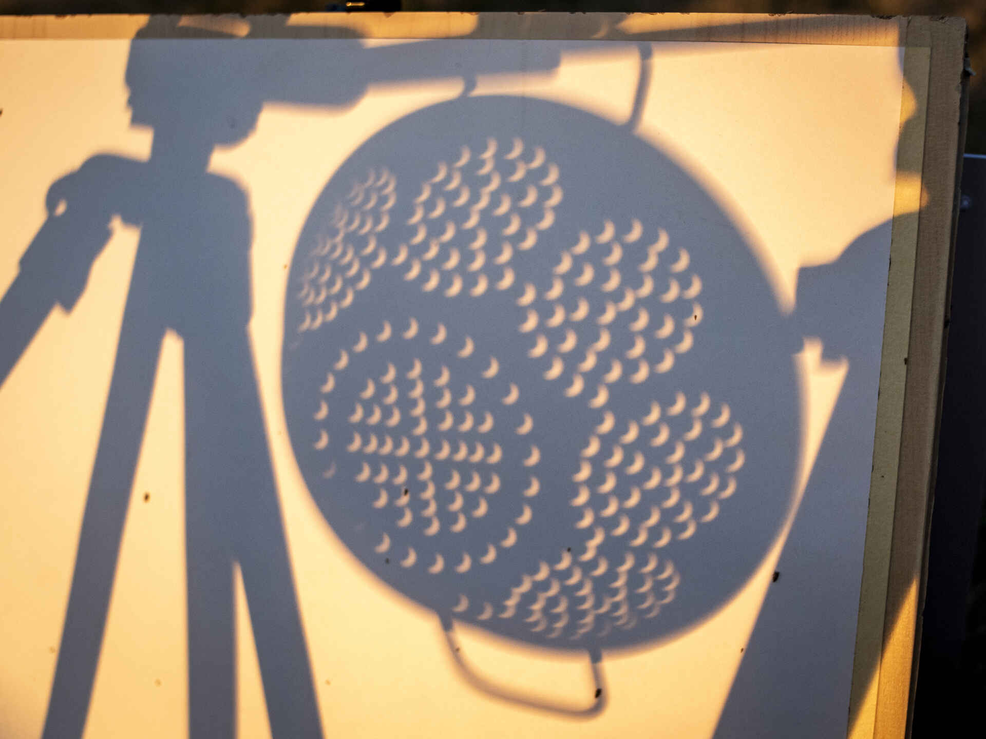 Farbbild (born digital): Sonnenlicht der teilweise bedeckten Sonne wird durch ein Nudelsieb auf einen weißen Karton projiziert.