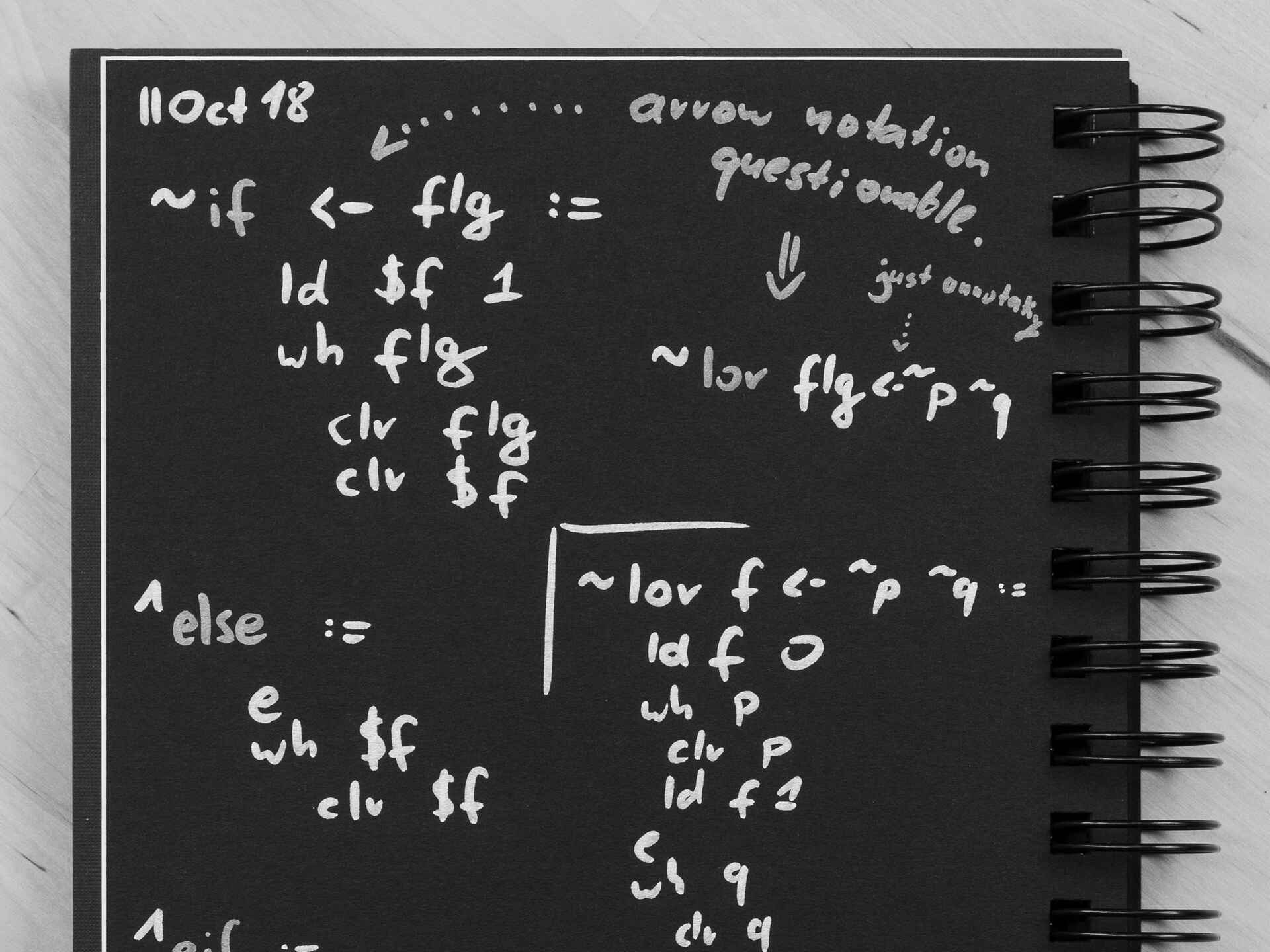 Schwarzweißbild, O⁠.⁠ ⁠i⁠.⁠ ⁠F⁠.: Seite eines Notizbuchs Notizen zum Entwurf eines Brainfuck-Compilers (siehe https://blog.jfrech.com/248) (Foto: Jonathan Frech, 9/2021)
