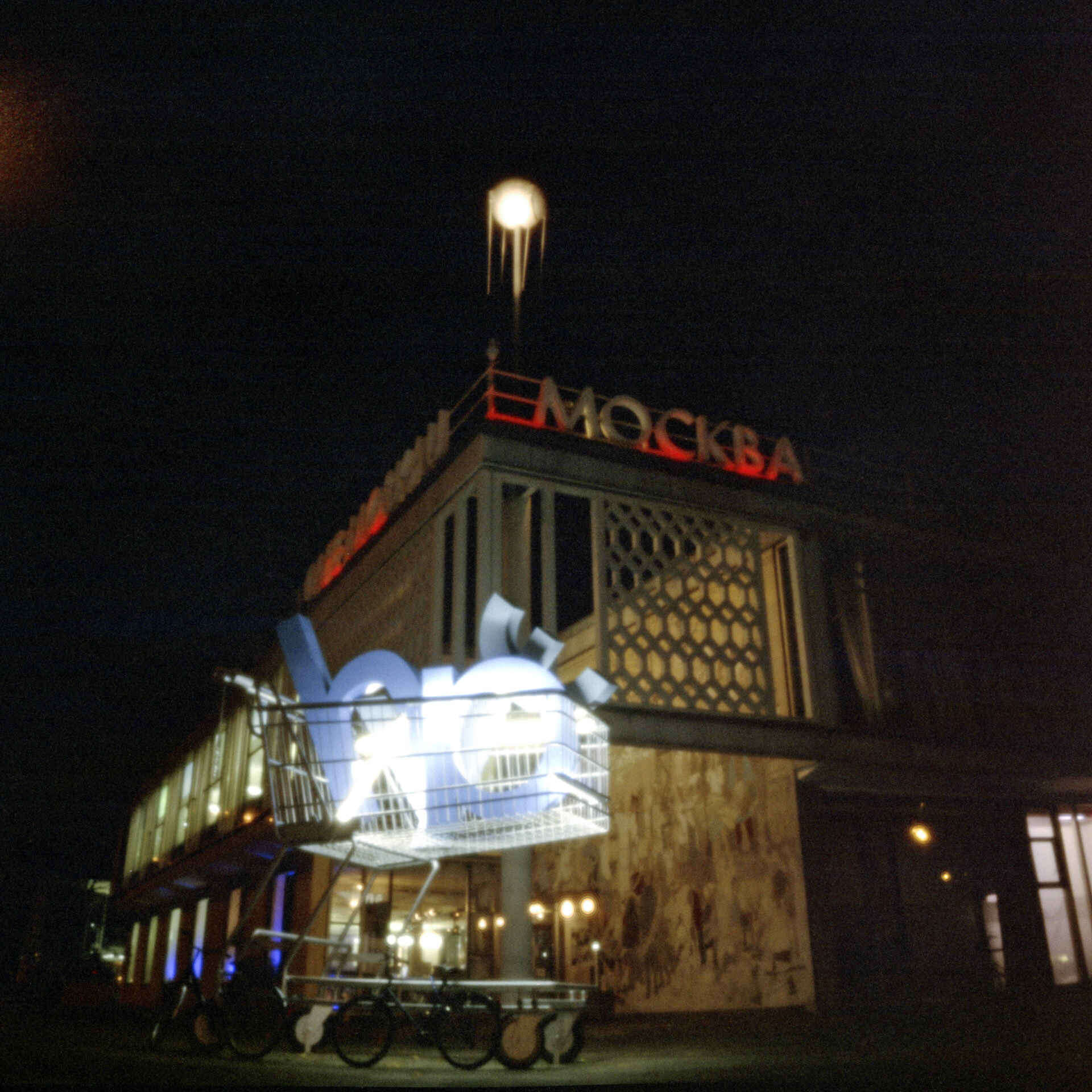 Lochkameraaufnahme (Farbbild): Außenaufnahme des Café Moskau in Berlin bei Nacht, davor steht ein überdimensionierter Einkausfwagen mit dem Hewlett-Packard-Logo. (Foto: Martin Frech, 11/2006)