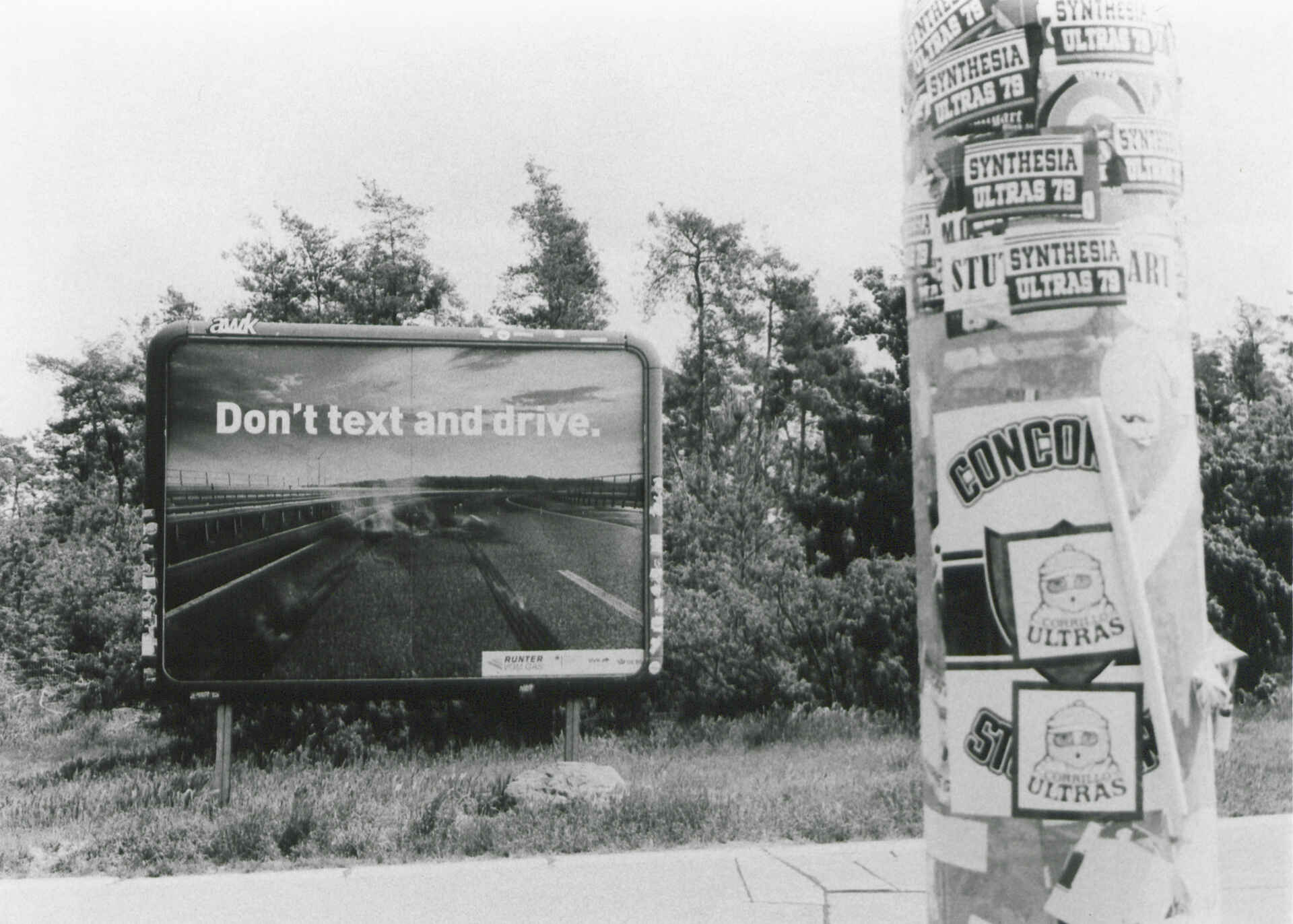 Unterwegs auf der Autobahn (Foma Ortho 400/Fomabrom Variant 111) (Foto: Martin Frech, 5/2025) Schwarzweißfoto: Das Bild zeigt ein Plakat mit der Aufschrift ›Don't text and drive‹. Im Vordergrund ein Pfosten mit verschiedenen Aufklebern u.a. ›SYNTHESIA ULTRAS 7‹ und ›CONCORDIA‹. (Foto: Martin Frech, 5/2025)
