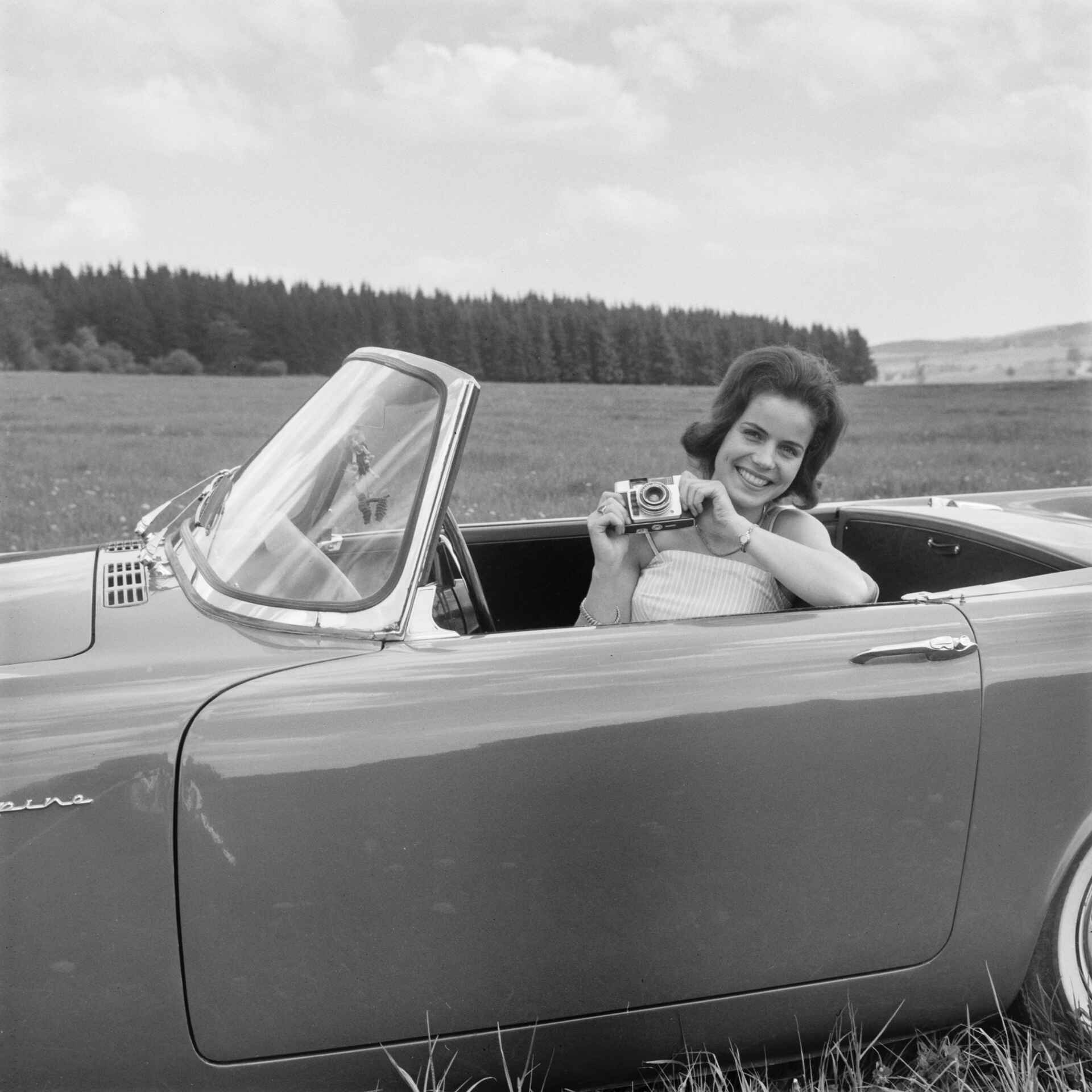 Freude über ein gelungenes Foto. (Foto: StadtA Rt./Näher, 6/1962) Schwarzweißfoto: Eine Frau sitzt mit ihrer Dacora-matic 4D hinter dem Lenkrad eines Sunbeam Alpine und freut sich. (Foto: StadtA Rt./Näher, 6/1962)