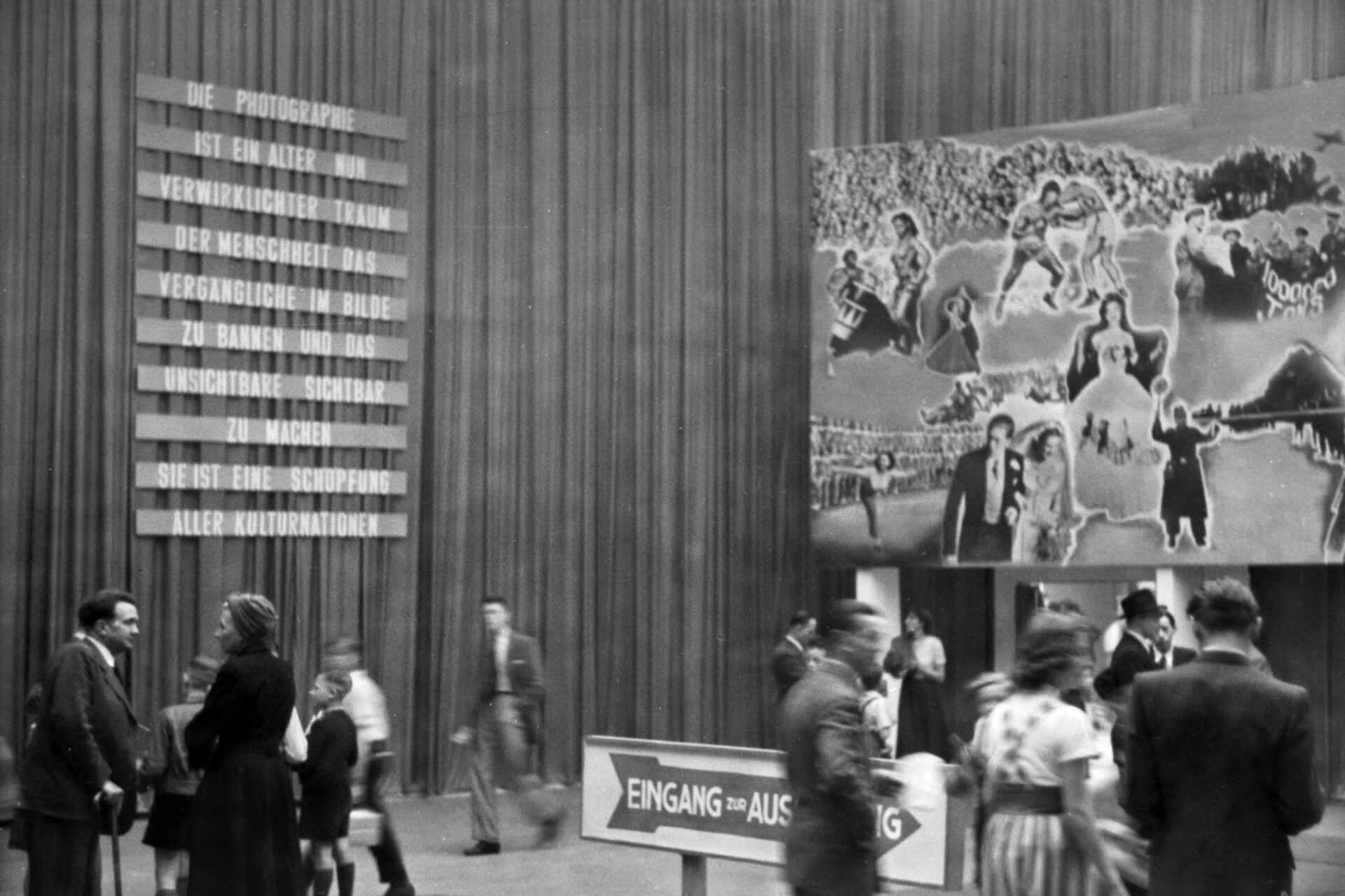 Schwarzweißbild (DSLR-Scan vom Negativ): Vorraum einer Bilderschau auf der ersten photokina (Photo- und Kino-Ausstellung in Köln). An der Wand steht: ›Die Photographie ist ein Alter, nun verwirklichter Traum der Menschheit, das Vergängliche im Bilde zu bannen und das Unsichtbare sichtbar zu machen. Sie ist eine Schöpfung aller Kulturnationen‹ (Foto: StadtA Rt./Dohm, 5/1950)