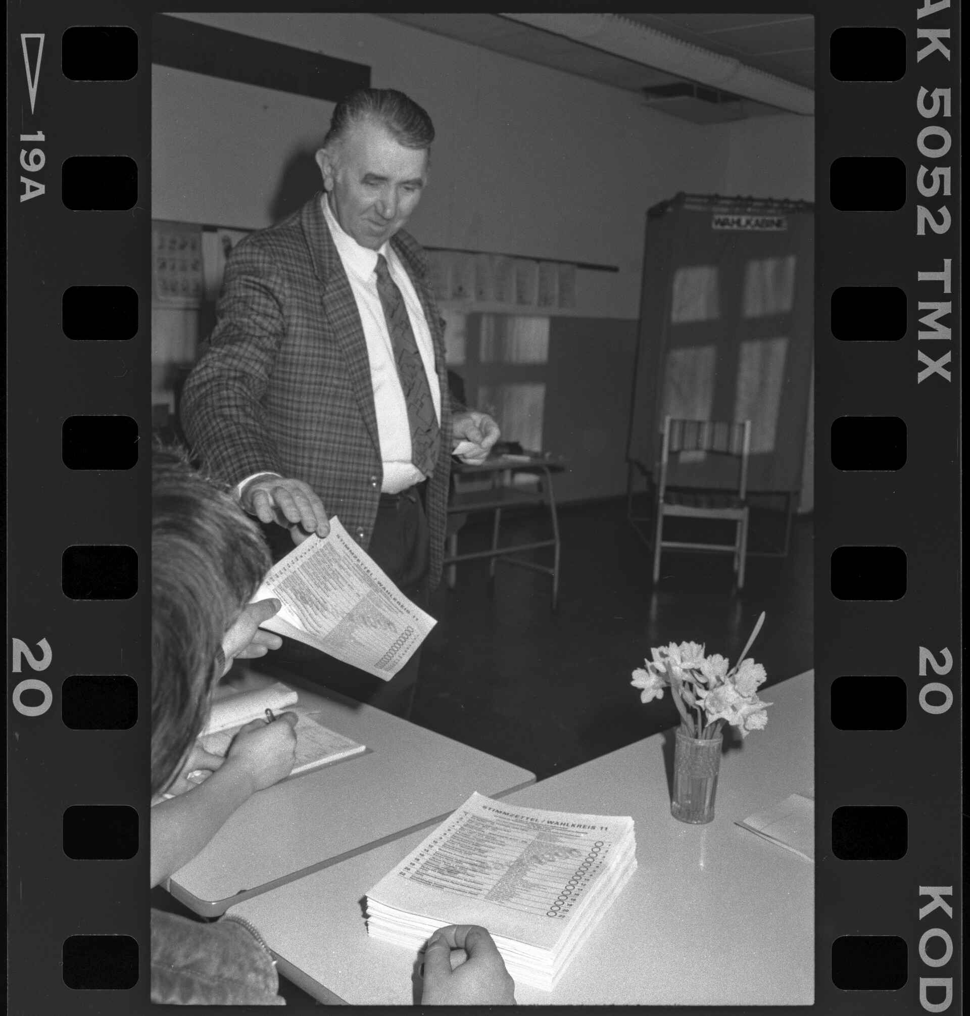 Wahllokal in Waren (Müritz); aus meinem Langzeit-Projekt ›Die letzten Male‹. (Foto: Martin Frech, 3/1990) Schwarzweißbild (Digitalisat vom Kleinbildnegativ): Ein Mann bekommt in einem DDR-Wahllokal einen Stimmzettel zur Volkskammerwahl 1990; im Hintergrund ist die Wahlkabine zu erkennen. (Foto: Martin Frech, 3/1990)