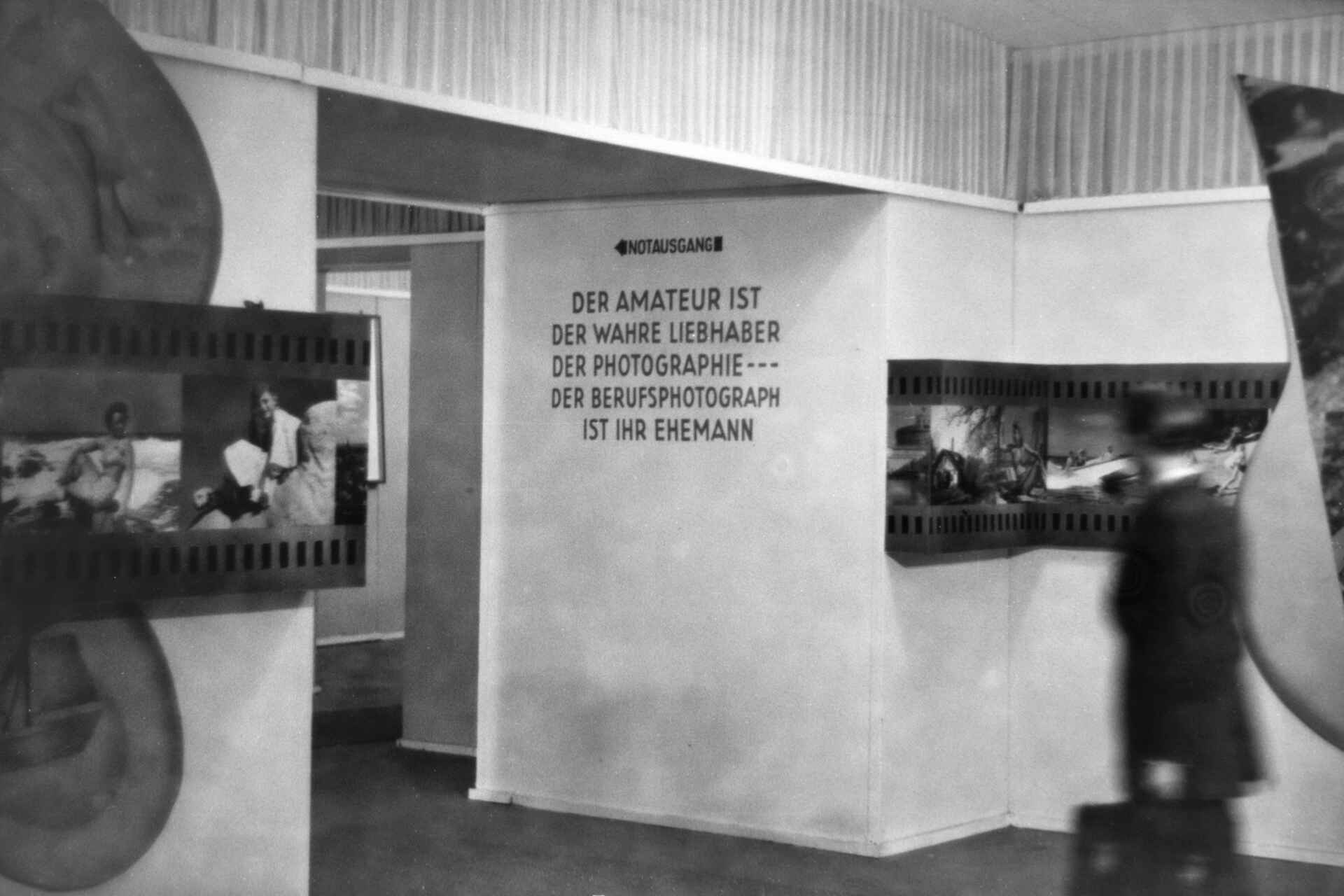 Schwarzweißbild (DSLR-Scan vom Negativ): Raum einer Bilderschau auf der ersten photokina (Photo- und Kino-Ausstellung in Köln). An der Wand steht: ›Der Amateur ist der wahre Liebhaber der Photographie --- der Berufsphotograph ist ihr Ehemann.‹ (Foto: StadtA Rt./Dohm, 5/1950)