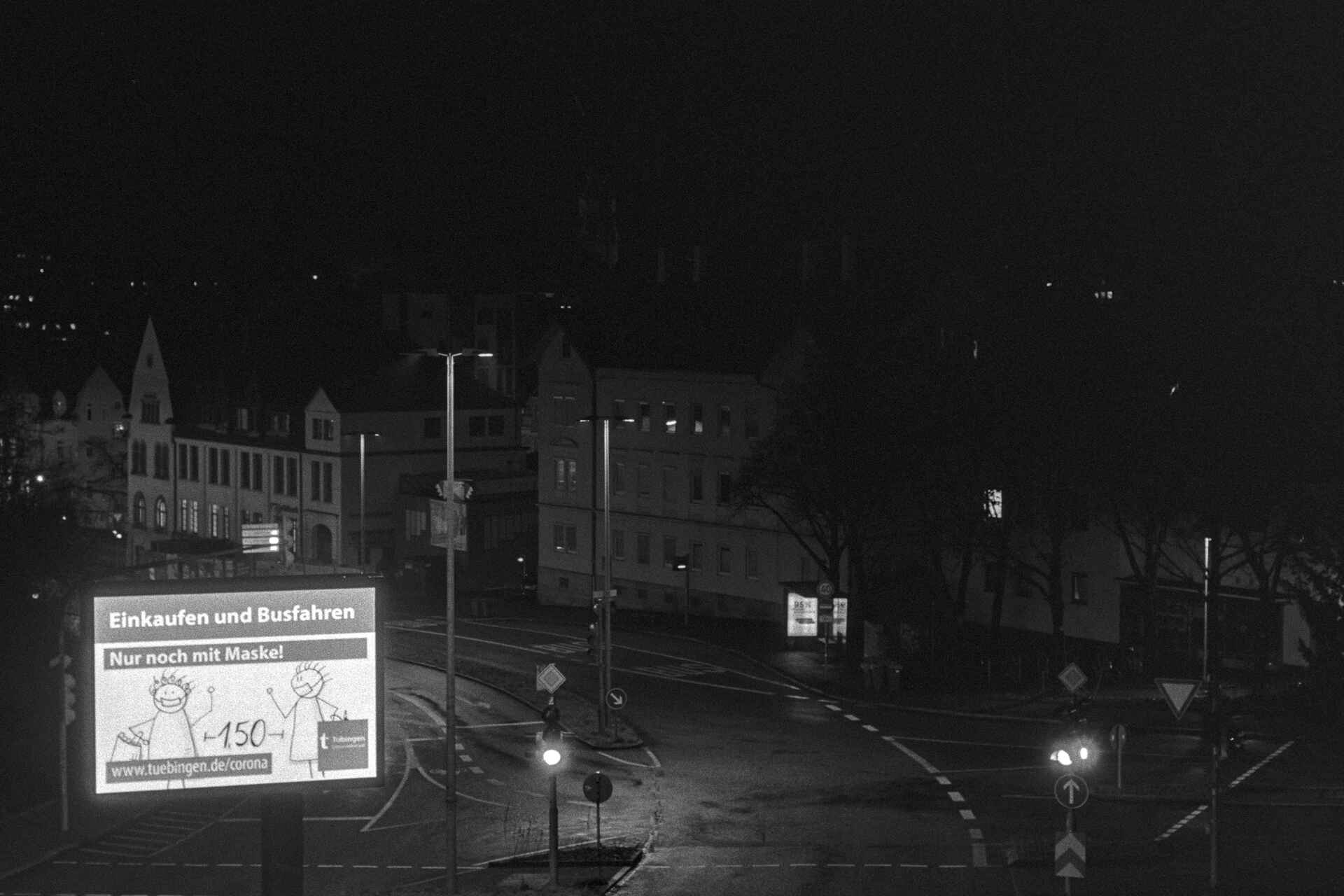 Schwarzweißbild (Scan vom Ilford-HP5+-Negativ): Werbung für die Corona-Regeln auf einer selbstleuchtenden Werbetafel in Tübingen. (Foto: Martin Frech, 1/2021)