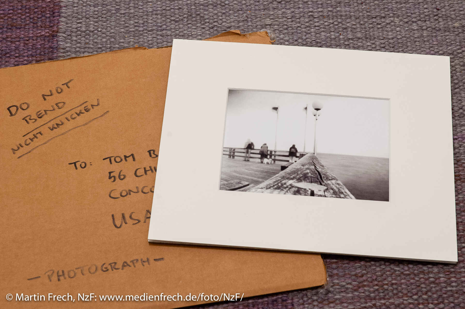 Ein Schwarzweißbild der Binzer Seebrücke des Fotografen Martin Frech im Passepartout; es liegt auf dem Boden auf einem Teppich und verdeckt den darunterliegenden Briefumschlag teilweise. (Foto: Martin Frech, 3/2007)