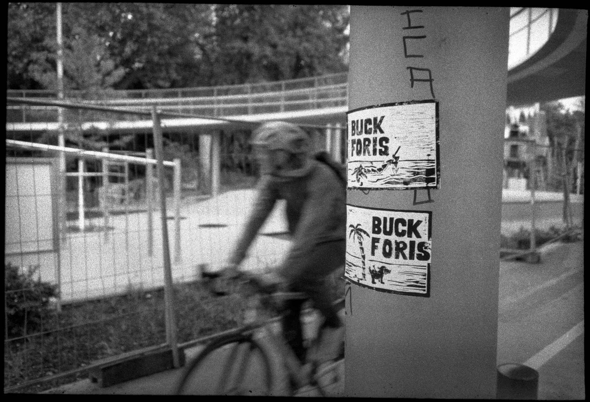 unter einer Fahrradbrücke in Tübingen (Foto: Martin Frech, 5/2026) Schwarzweiß (DSLR-Scan vom Kleinbild-Negativ): Zettel am Brückenpfeiler einer Fahrradbrücke in Tübingen: ›Buck Foris‹; wahrscheinlich ein Bezug zum Oberbürgermeister Boris Palmer. Im Hintergrund bewegungsunscharf ein Fahrradfahrer.