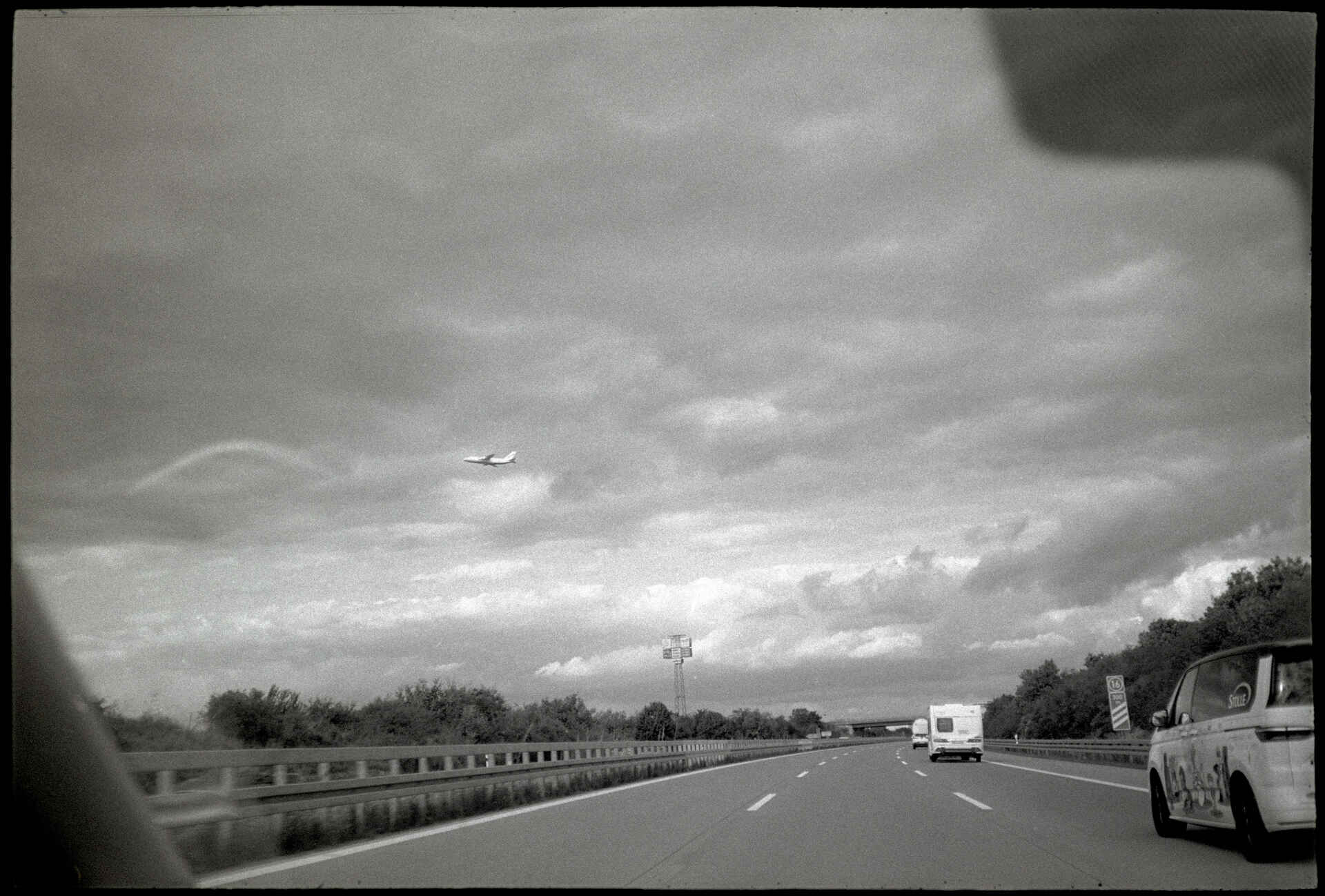 Autobahn A9 bei Leipzig (Foto: Martin Frech, 9/2026) Schwarzweißbild (digitalisiert vom Kodak-Tri-X-Kleinbild-Negativ): Blick aus dem fahrenden Pkw auf die Autobahn A9 bei Leipzig; am Himmel fliegt ein Flugzeug.