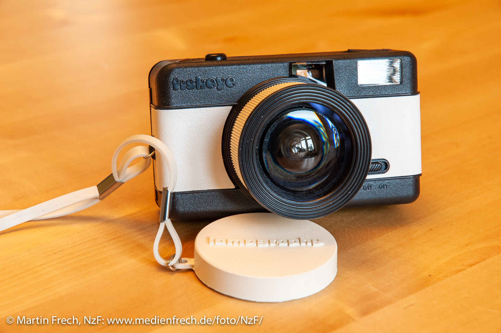 Die Lomo-Fischauge-Kamera ›Lomo Fisheye [One]‹ in weiß mit Objektivkappe auf einer Holzplatte.