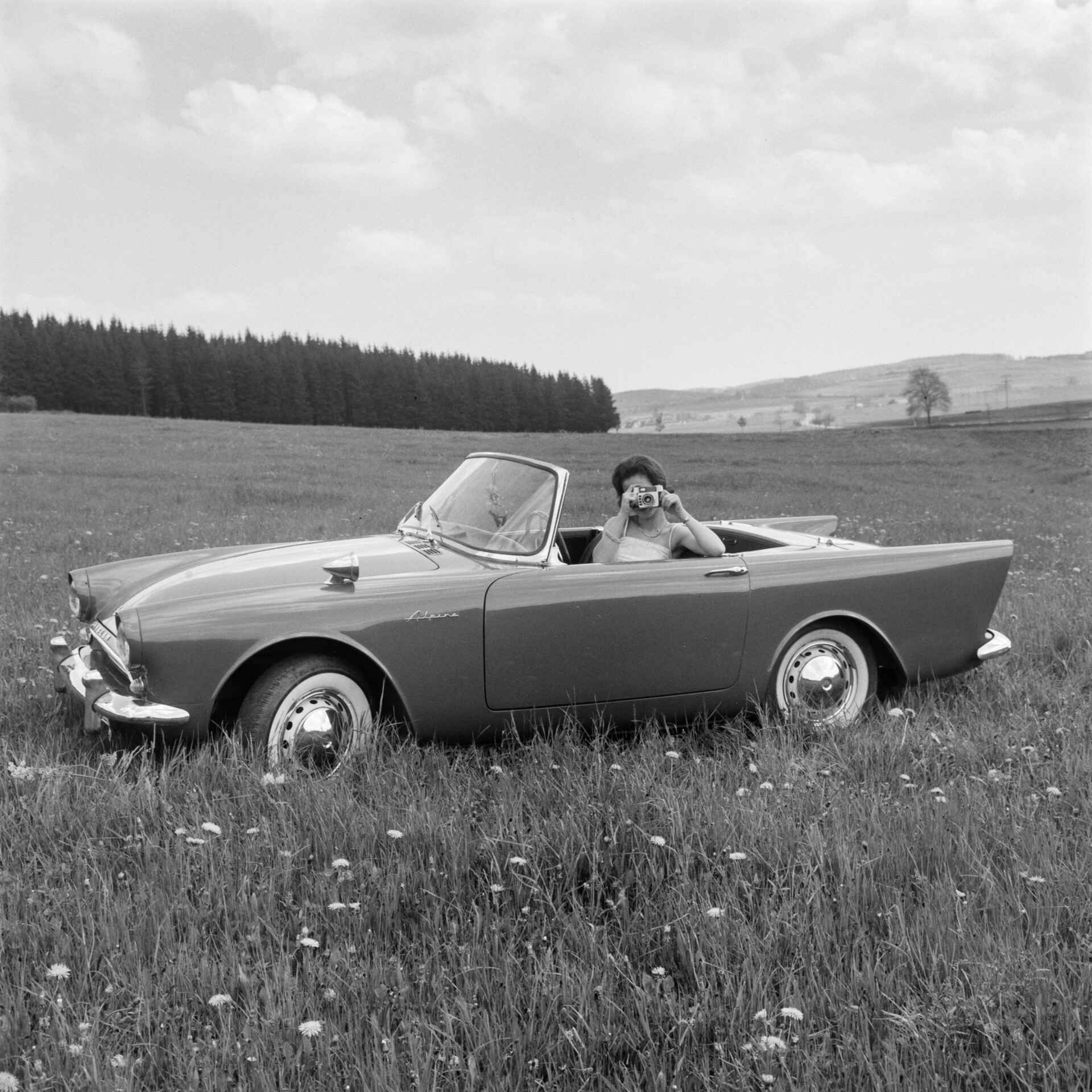 Spielt auch im ersten James-Bond-Film eine Rolle: der Sunbeam Alpine. (Foto: StadtA Rt./Näher, 6/1962) Schwarzweißfoto: Eine Frau sitzt hinter dem Lenkrad eines Sunbeam Alpine und fotografiert. (Foto: StadtA Rt./Näher, 6/1962)