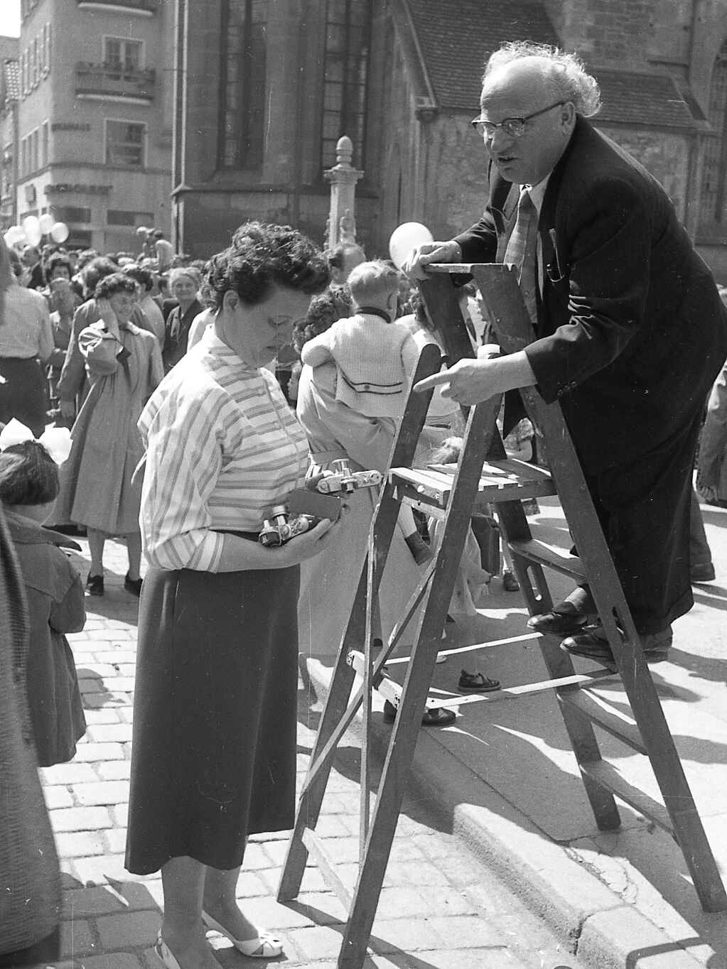 Peter Dohm wählt eine Leica (Foto: StadtA Rt./Dohm, 1956) Schwarzweißbild: Ein Fotograf steht auf einer Leiter und lässt sich von seine Assistentin eine Kamera reichen. (Foto: StadtA Rt./Dohm, 1956)