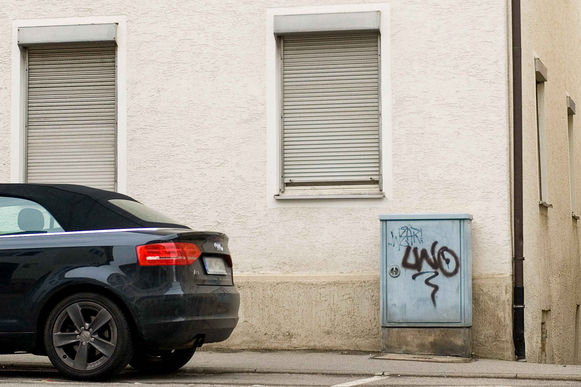 Detailansicht einer Hausecke (Erdgeschoss), die Rolläden sind heruntergelassen; davor ein grauer Anschlusskasten der Telekom mit Graffiti ›LWO‹. Auf dem Parkstreifen parkt ein schwarzes Auto. (Foto: Martin Frech, 5/2010)