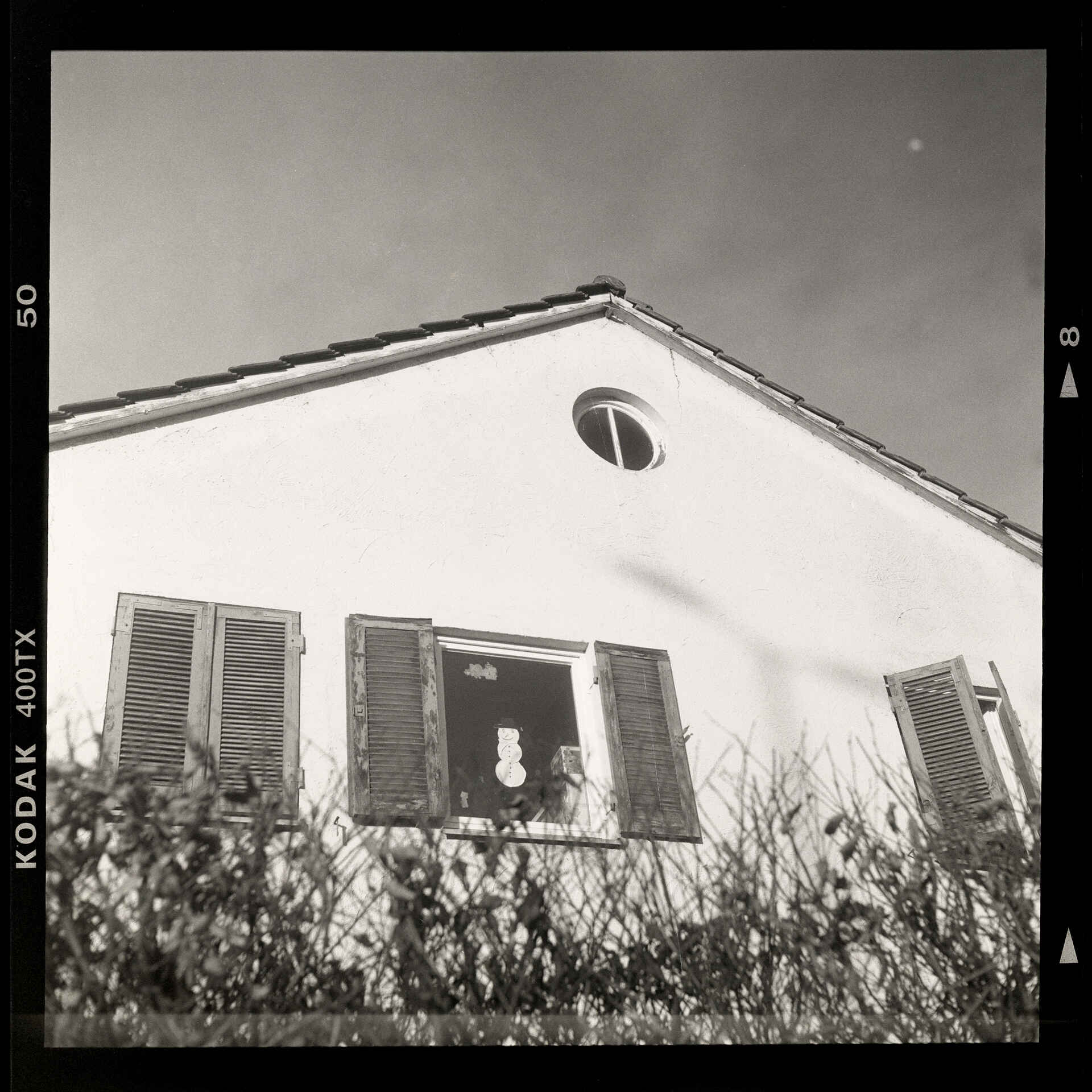 Fenster mit Schneemann; Rolleiflex 3.5, Gelbfilter, Kodak Tri-X, Kodak Xtol 1+1 (Foto: Martin Frech, 2025) Schwarzweißbild: Giebelwand eines Wohnhauses in Untersicht. Fenster mit Fensterläden, teilw. offen. In einem Fenster hängt ein Papier-Schneemann. (Foto: Martin Frech, 2025)