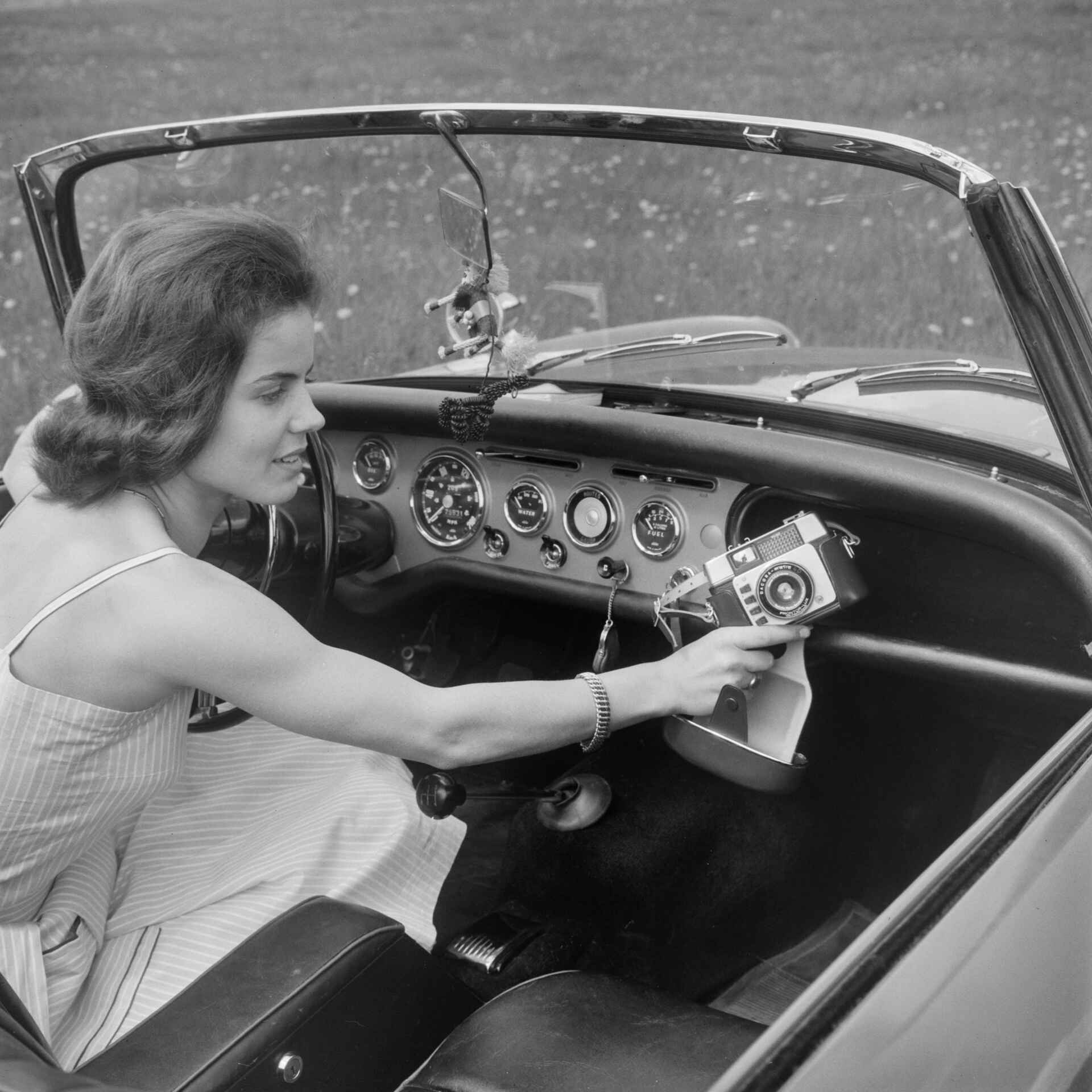 Die Dacora-matic passt so gar nicht zur beeindruckenden Uhrensammlung am Armaturenbrett des Sunbeams. (Foto: StadtA Rt./Näher, 6/1962) Schwarzweißfoto: Eine Frau sitzt hinter dem Lenkrad eines Sunbeam Alpine und holt eine Dacora-matic 4D aus dem Handschuhfach. (Foto: StadtA Rt./Näher, 6/1962)