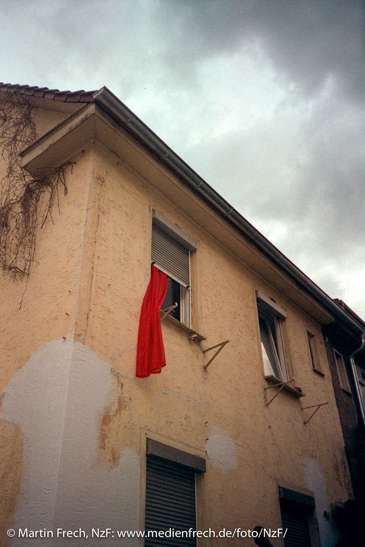 Farbbild: Haus in Untersicht, aus einem Fenster hängt ein roter Vorhang. Der Himmel ist dunkel bewölkt. (Foto: Martin Frech, 7/2017)