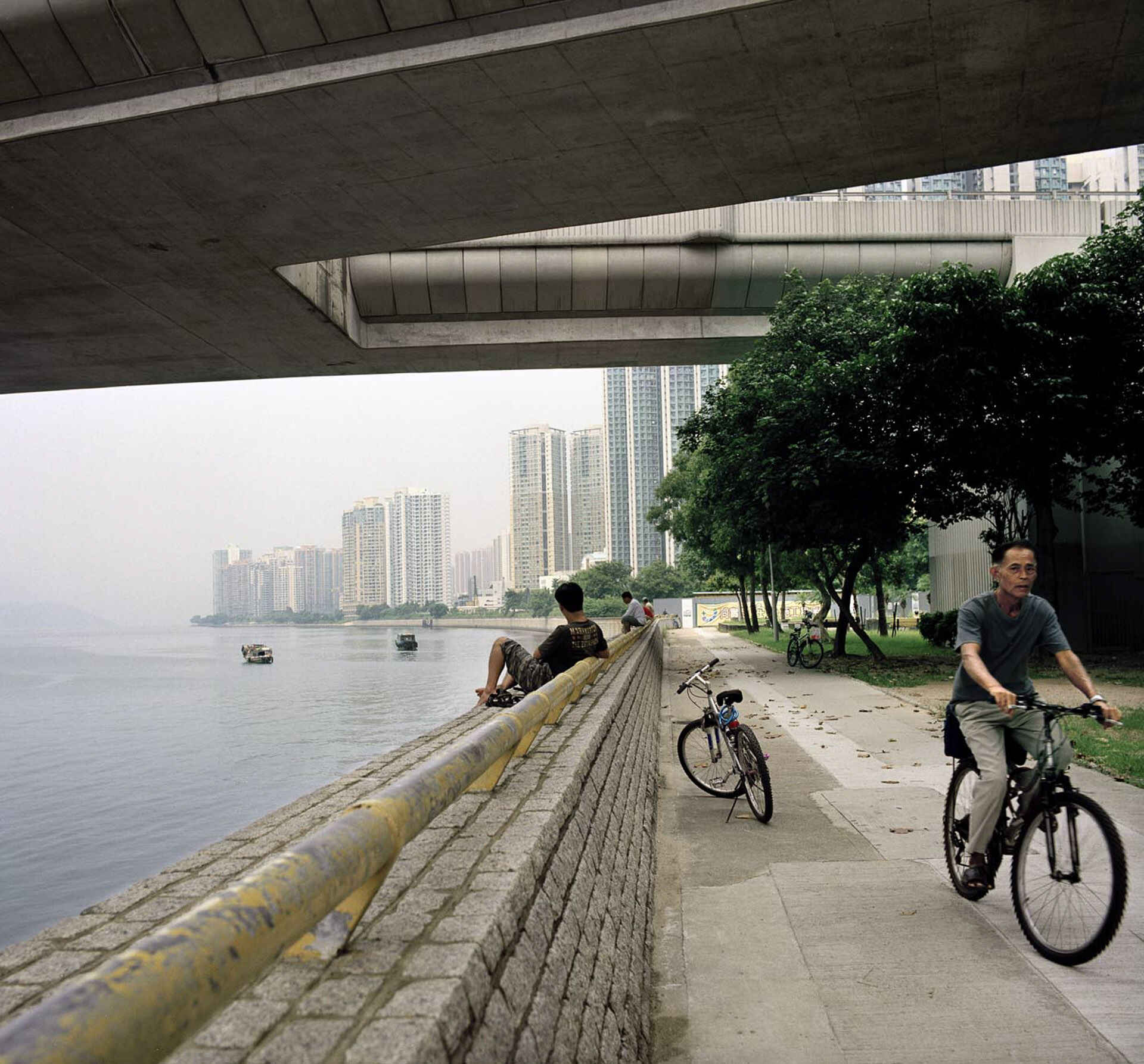 Tobias Vollmer: aus »China Construction« (Foto: Tobias Vollmer, 2012) Farbbild: Befestigtes Flussufer in China unter einer Brücke, rechts ein Radfahrer, im Hintergrund Hochhäuser. (Foto: Tobias Vollmer, 2012)