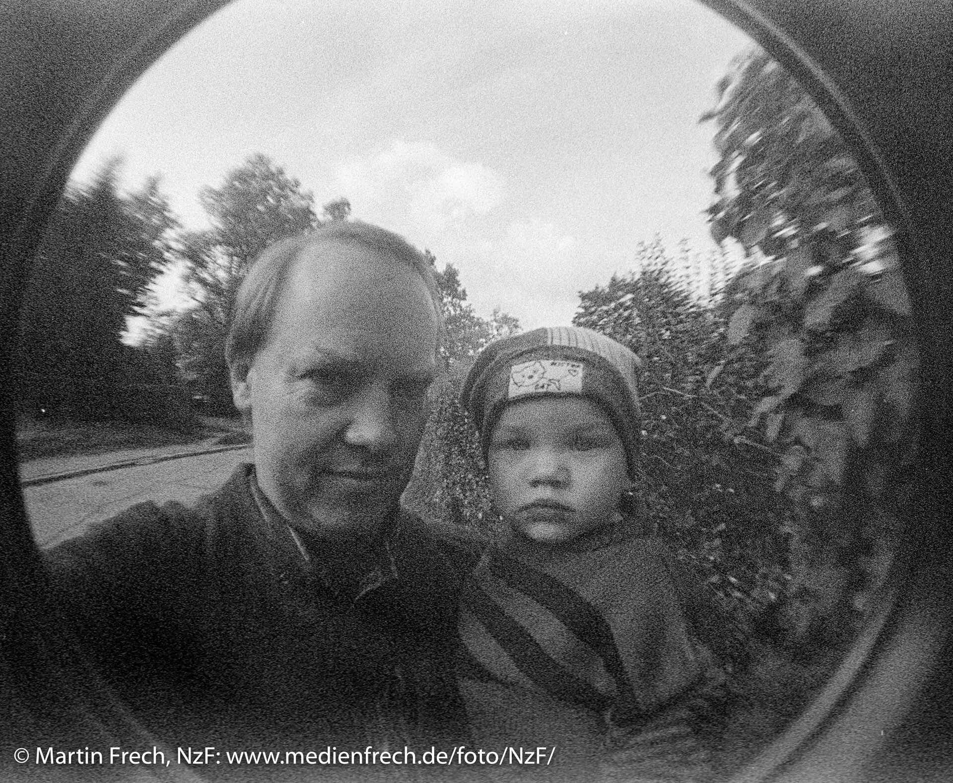 Fischaugen-Aufnahme mit der ›Lomo Fisheye [One]‹, schwarzweiß: Selfie eines Mannes mit Kind auf dem Arm.