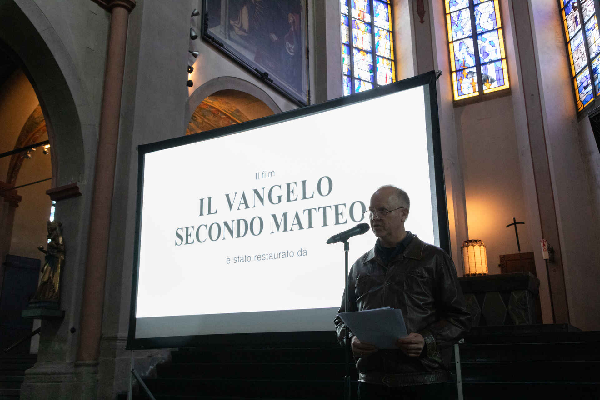 Farbbild: Martin Frech am Mikrophon in der Kirche St. Maria in Lyskirchen, Köln vor einer Leinwand mit dem Filmtitel ›Il Vangelo secondo Matteo‹. Im Hintergrund bunte Kirchenfenster. (Foto: Tobias D. Kern, 4/2025)