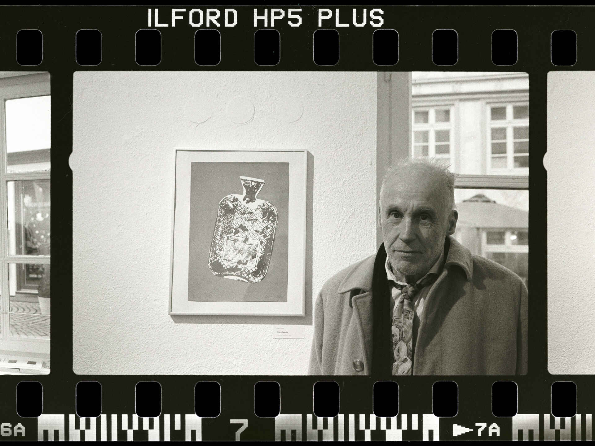 Schwarzweißbild: Portrait des Künstlers Dieter Löchle neben seinem Werk ›Wärmflasche‹ in der Jahresausstellung des Künstlerbunds Tübingen. (Foto: Martin Frech, 1/2025)