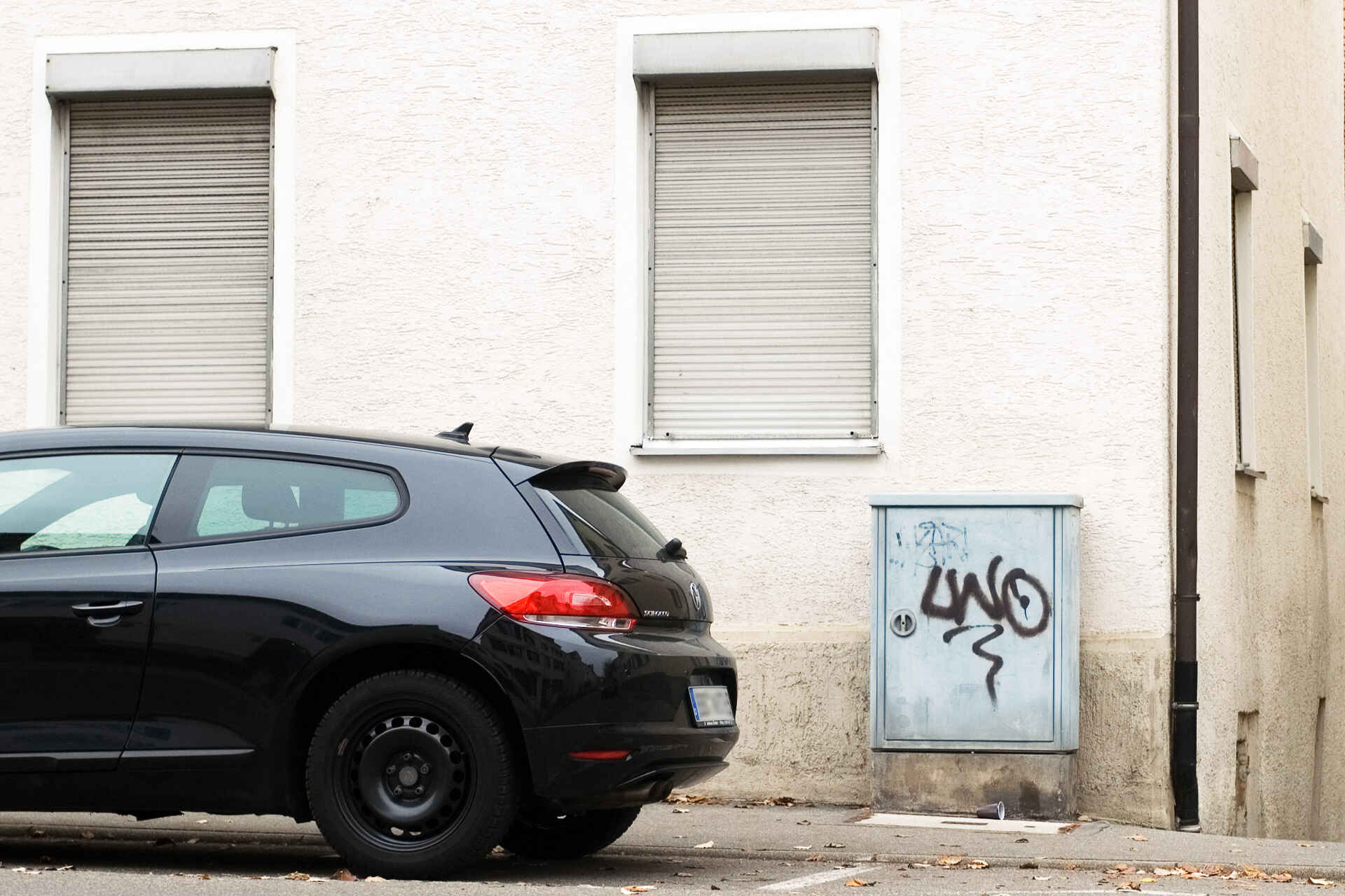 Detailansicht einer Hausecke (Erdgeschoss), die Rolläden sind heruntergelassen; davor ein grauer Anschlusskasten der Telekom mit Graffiti ›LWO‹. Auf dem Parkstreifen parkt ein schwarzes Auto. (Foto: Martin Frech, 11/2011)