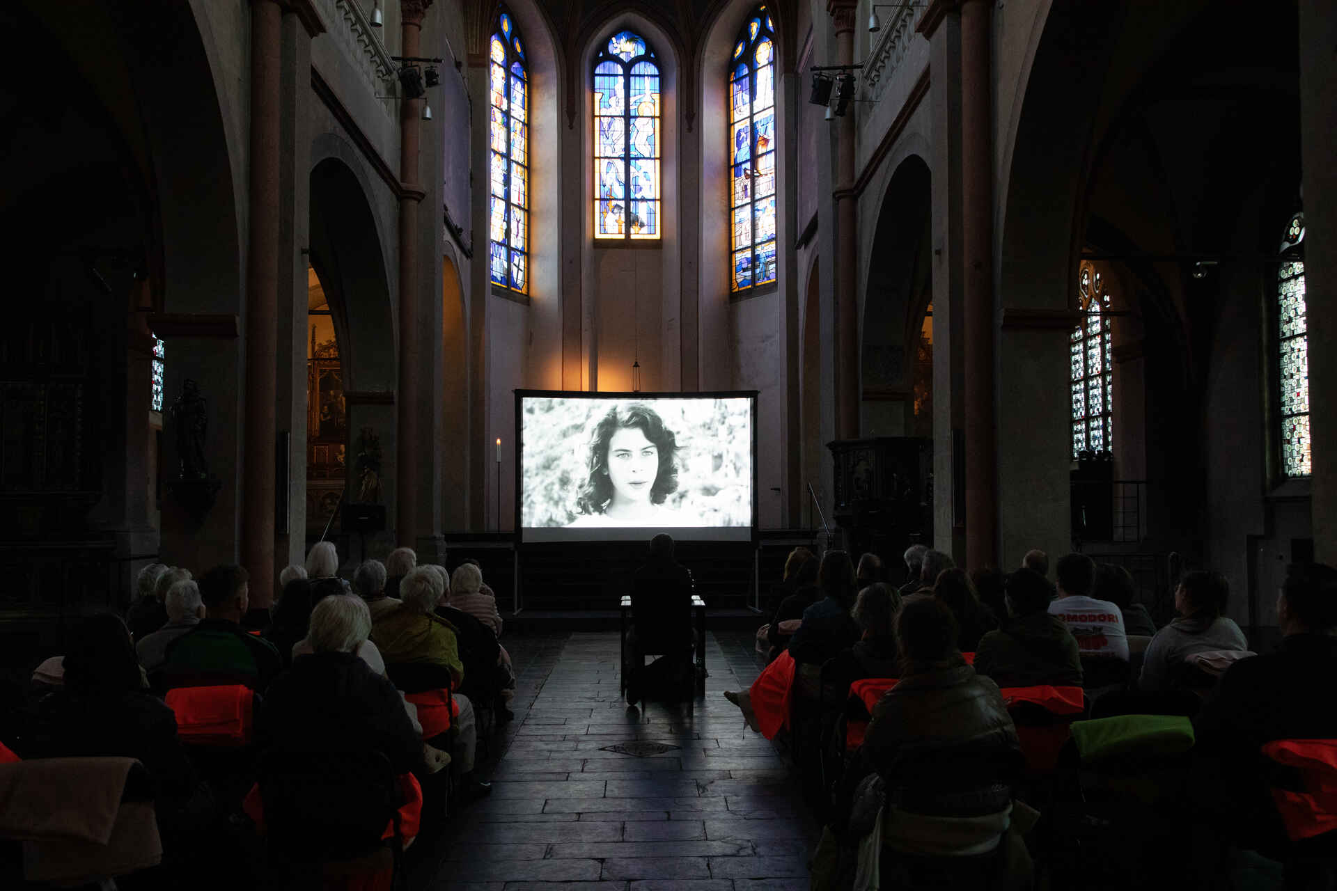 Farbbild: Filmvorführung in der Kirche St. Maria in Lyskirchen, Köln. Im Hintergrund bunte Kirchenfenster. (Foto: Tobias D. Kern, 4/2025)