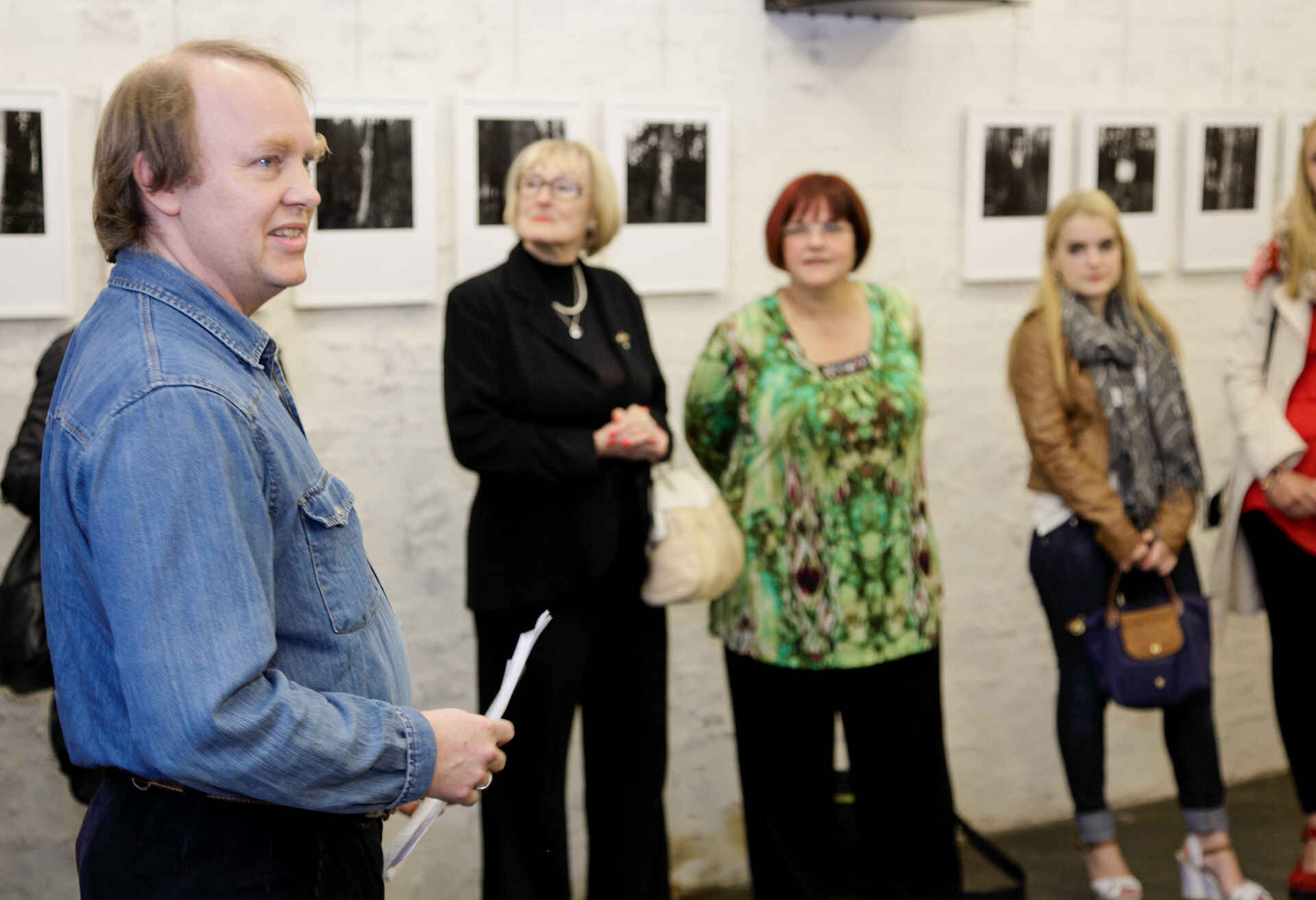 Martin Frech redet zur Eröffnung der Ausstellung ›Stigmata‹ in der schaelpic photokunstbar, Köln. (Foto: Andrea Otto, 9/2012) Farbbild: Redner im Vordergrund, Publikum unscharf im Hintergrund; an der Wand unscharf Schwarzweißfotos unter Passepartout in weißen Bilderrahmen. (Foto: Andrea Otto, 9/2012)