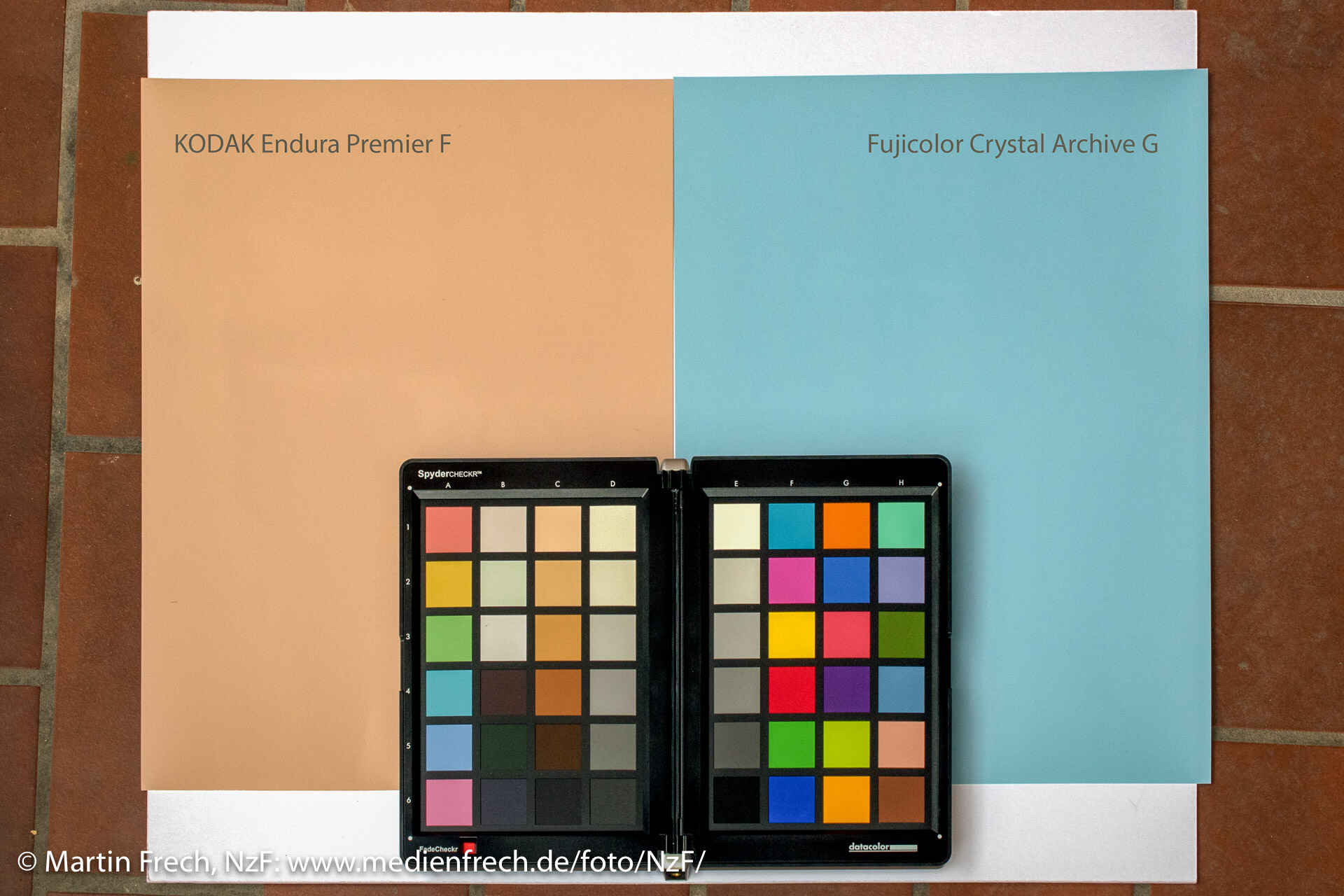 Die Papiere von Kodak und Fuji haben vor der Entwicklung unterschiedlich gefärbte Schichten. (Foto: Martin Frech, 5/2017) Auf einem gefliesten Boden liegen je ein nicht entwickelter Bogen Kodak Endura Premier F und Fujifilm Fujicolor Crystal Archive G nebeneinander auf einem weißen Blatt Papier. Darauf liegt die aufgeklappte ColorChecker-Testtafel.
