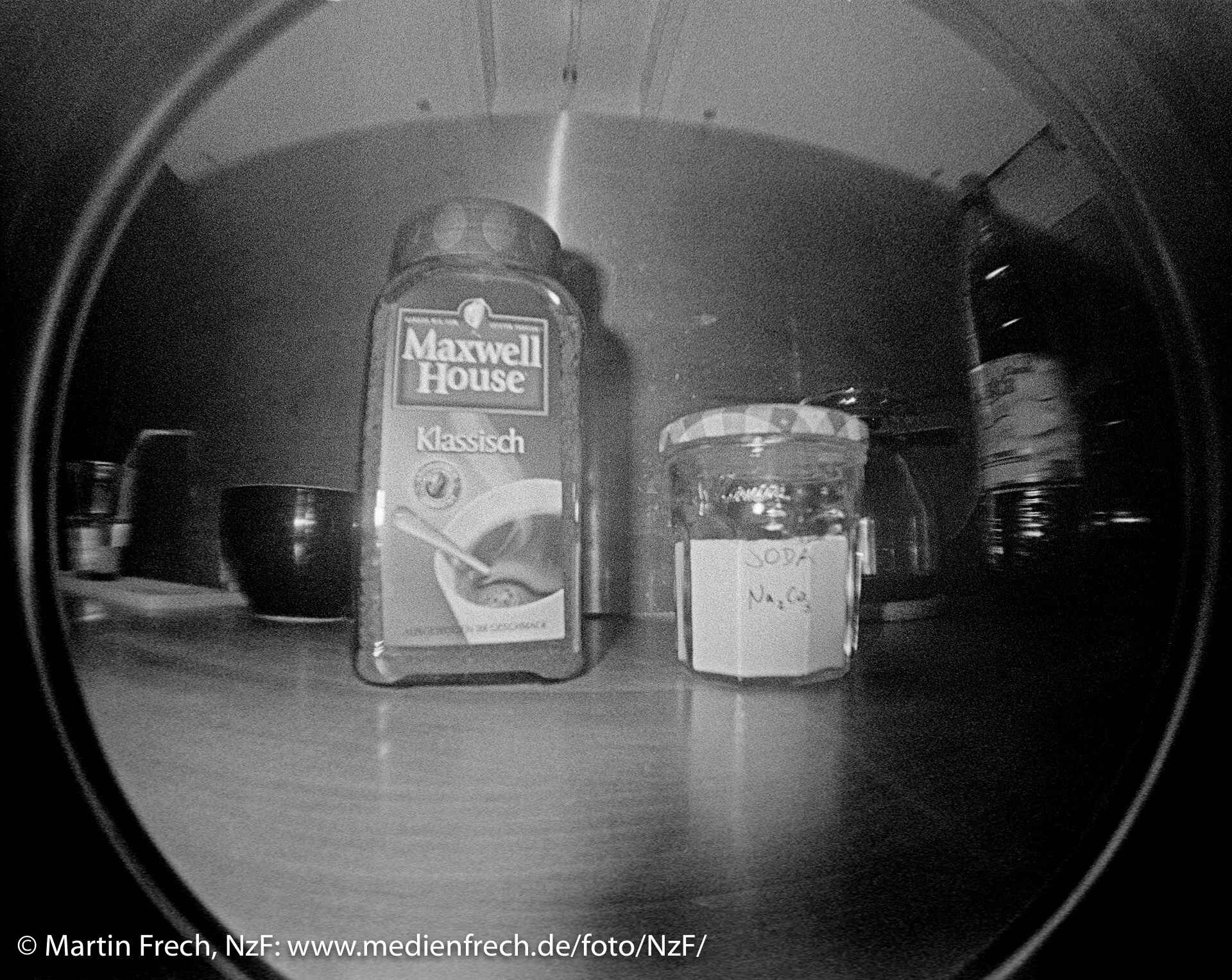 Fischaugen-Aufnahme mit der ›Lomo Fisheye [One]‹, schwarzweiß, direktes Blitzlicht: Verpackung mit Instant-Kaffeepulver (Etikett: Maxwell House­ Klassisch) und ein Schraubglas mit Soda (beschriftet: Soda Na₂CO₃) auf einer Küchenarbeitsplatte.
