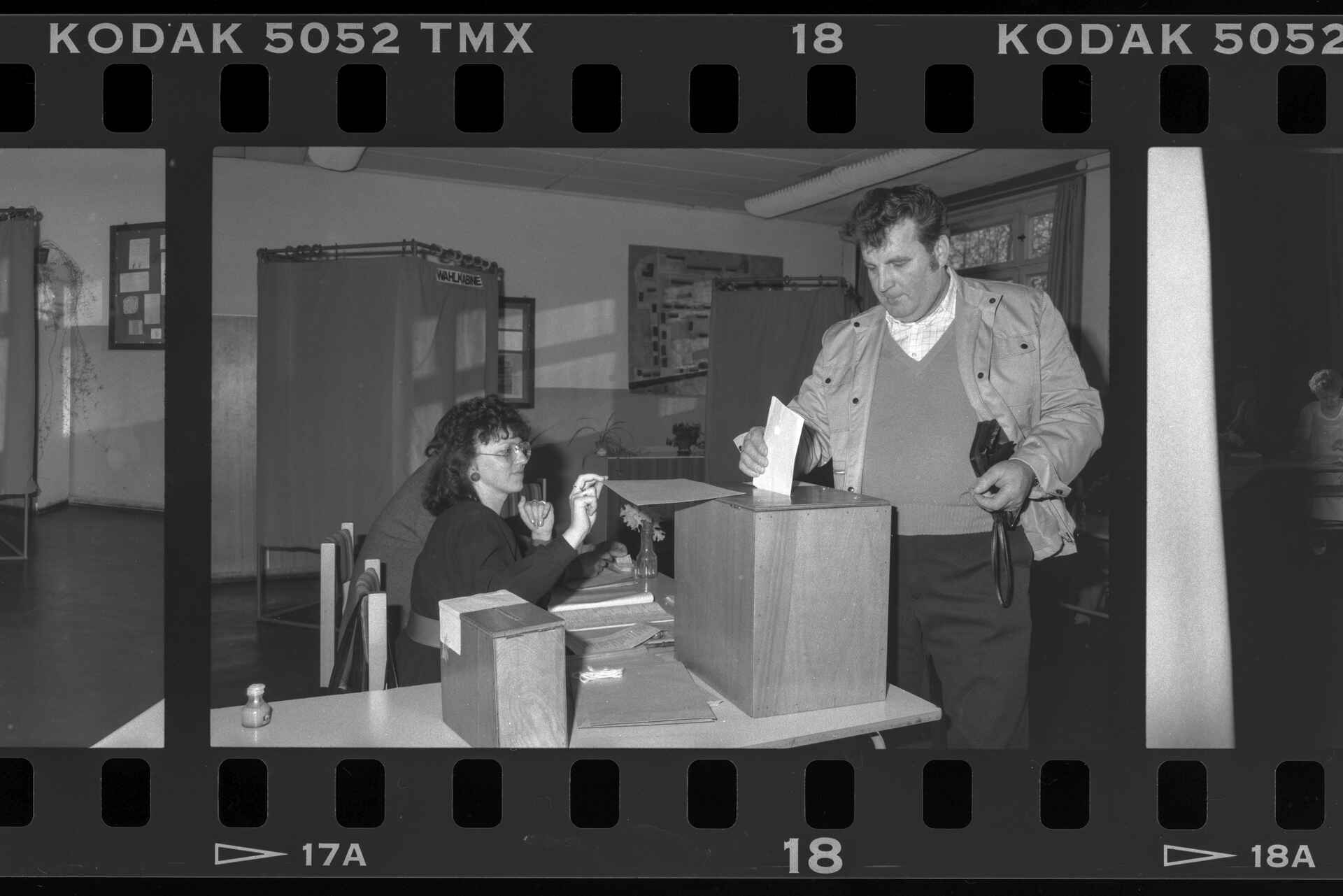 Schwarzweißbild (Digitalisat vom Kleinbildnegativ): Ein Mann gibt in einem DDR-Wahllokal seinen Stimmzettel zur Volkskammerwahl 1990 in die Wahlurne; im Hintergrund ist die Wahlkabine zu erkennen. (Foto: Martin Frech, 3/1990)