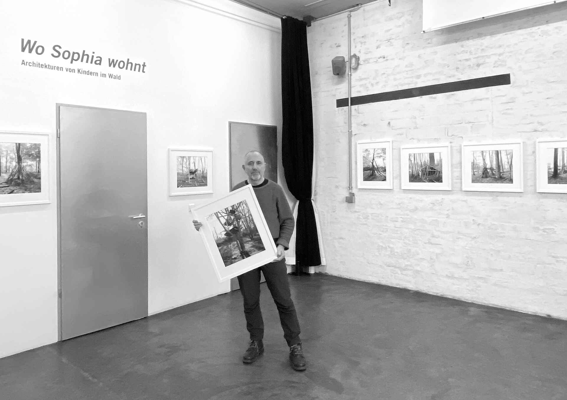 Schwarzweißbild: Der Fotograf Tobias D. Kern steht in seiner Ausstellung ›Wo Sophia wohnt‹ (schaelpic photokunstbar, Köln) und hält eine gerahmte Fotografie in der Hand. (Foto: Claudia Maas, 11/2024)