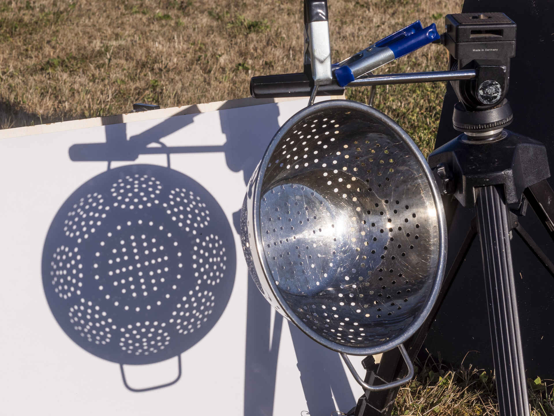 Die Löcher des Nudelsiebs wirken als Lochblendenstruktur: Vor Beginn der Bedeckung wird durch jedes Loch die kreisrunde Sonne abgebildet. Farbbild (born digital): Das Bild der Sonne wird durch ein Nudelsieb auf einen weißen Karton projiziert.